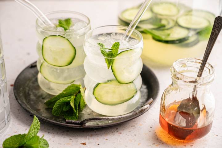 Ultra Hydrating Cucumber Water Two glasses of refreshing cucumber mint water with glass straws sit on a metal tray. A jar of honey and a bundle of fresh mint are nearby