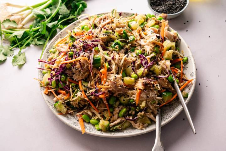 25 Rotisserie Chicken Recipes A vibrant Asian salad with shredded chicken, colorful cabbage, edamame, carrots, and cucumbers. Garnished with black sesame seeds. Fresh cilantro and green onions are nearby.