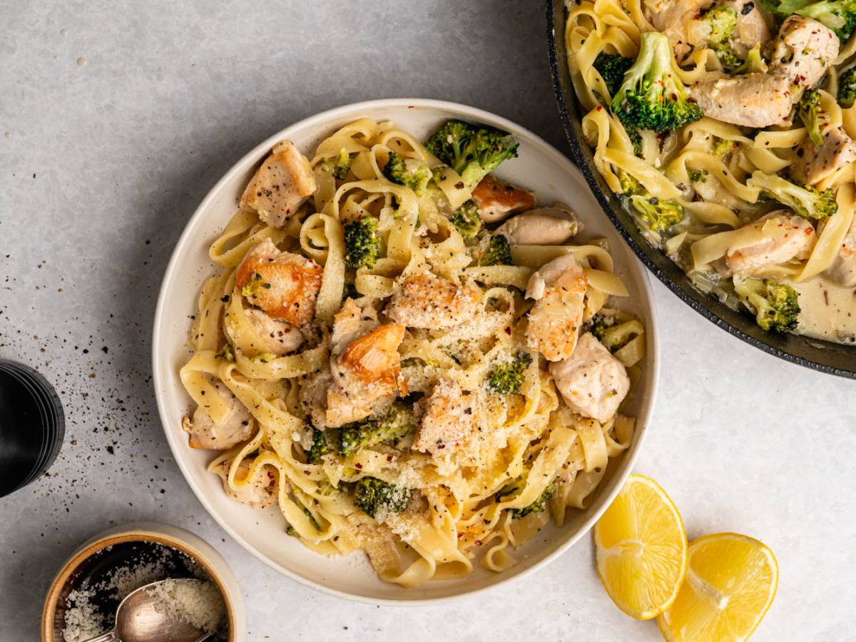 High-Protein Chicken Plate of creamy chicken and broccoli pasta topped with parmesan, next to a similar skillet. Lemon wedges and pepper grinder add fresh vibes.
