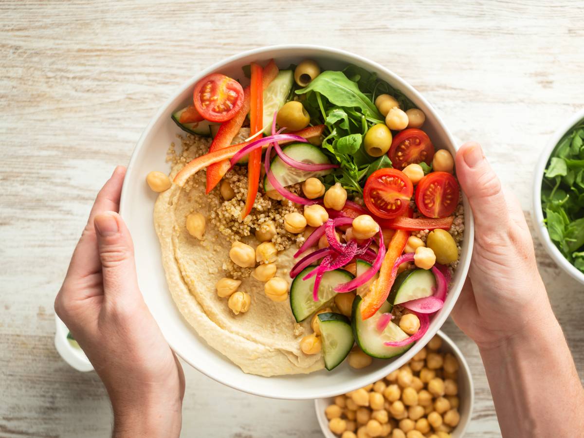 Gluten Free Woman hands holding a bowl with creamy hummus, quinoa, chickpeas, cucumbers, arugula, and red onions.