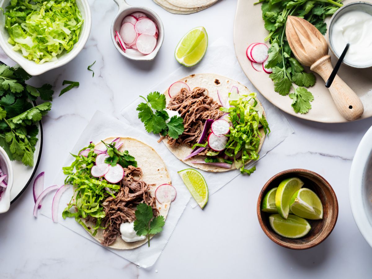 Mexican Beef Recipes Crockpot tri-tip shredded tacos with red onion, cilantro, lettuce, and radishes served on corn tortillas.