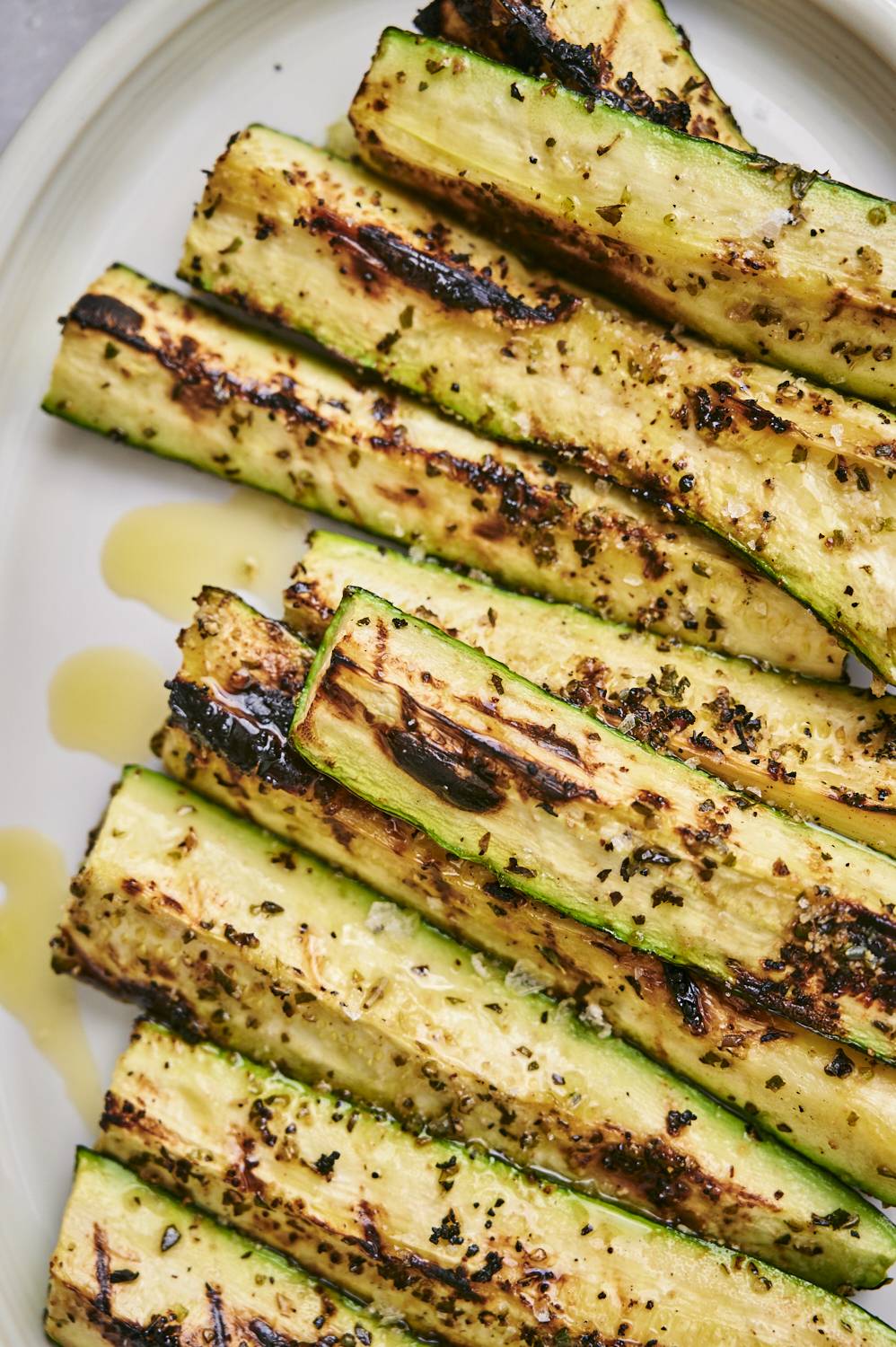 Zucchini spears with brown grill marks served on a white plate with olive oil, flake salt, and pepper.