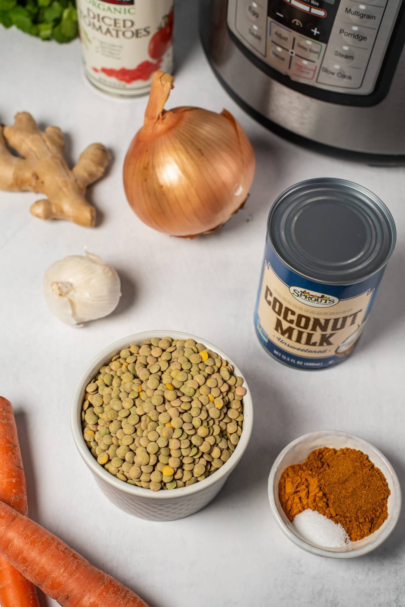 Ingredients for lentil coconut curry, including lentils, coconut milk, onion, garlic, ginger, carrots, and spices, displayed on a white surface.