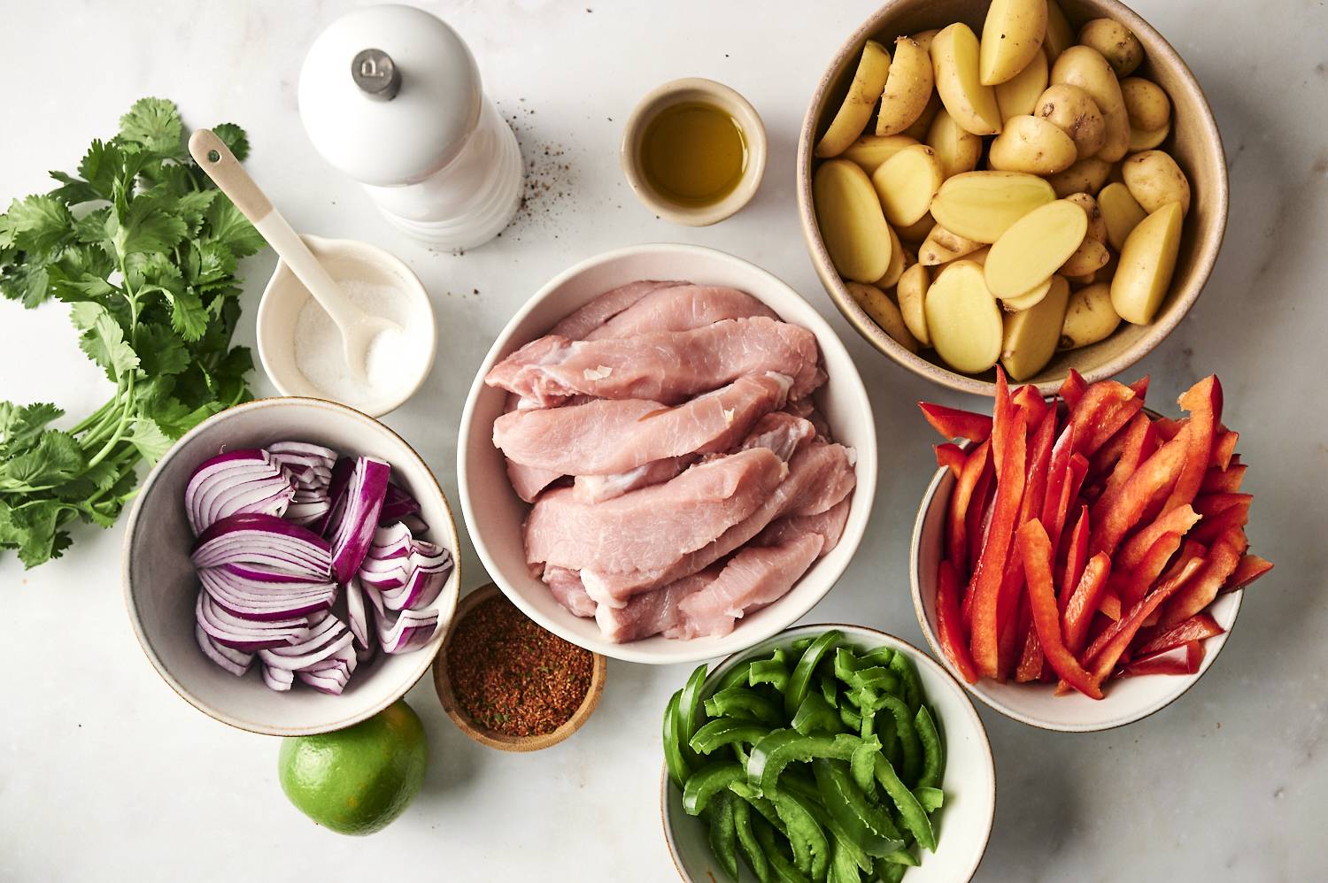 A flat lay of fresh ingredients including raw pork chops, sliced potatoes, red and green bell peppers, red onions, cilantro, lime, and spices, arranged on a white surface.