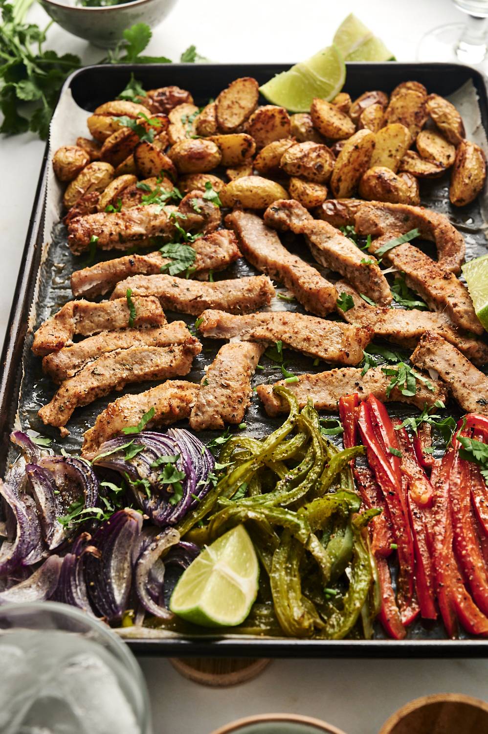 A sheet pan filled with roasted pork chop strips, golden-brown potatoes, sautéed bell peppers, and red onions, garnished with fresh cilantro and lime wedges.