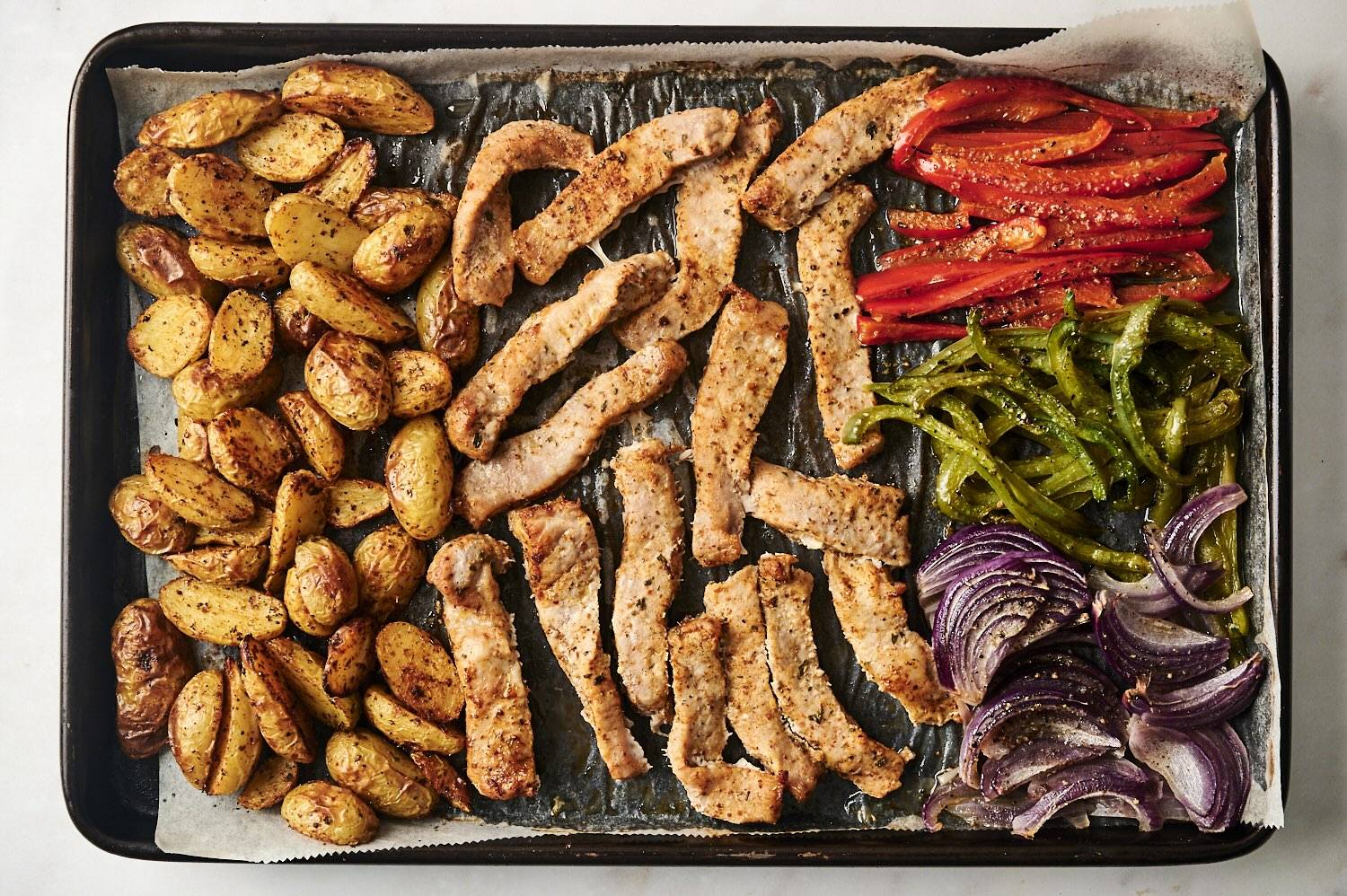 A sheet pan filled with roasted pork chop strips, seasoned baby potatoes, red and green bell peppers, and red onions, all cooked to perfection.