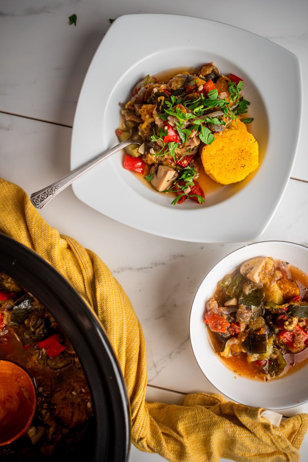 Crockpot ratatouille with stewed eggplant, zucchini, tomatoes, onions, and mushrooms in a bowl with polenta on the side.