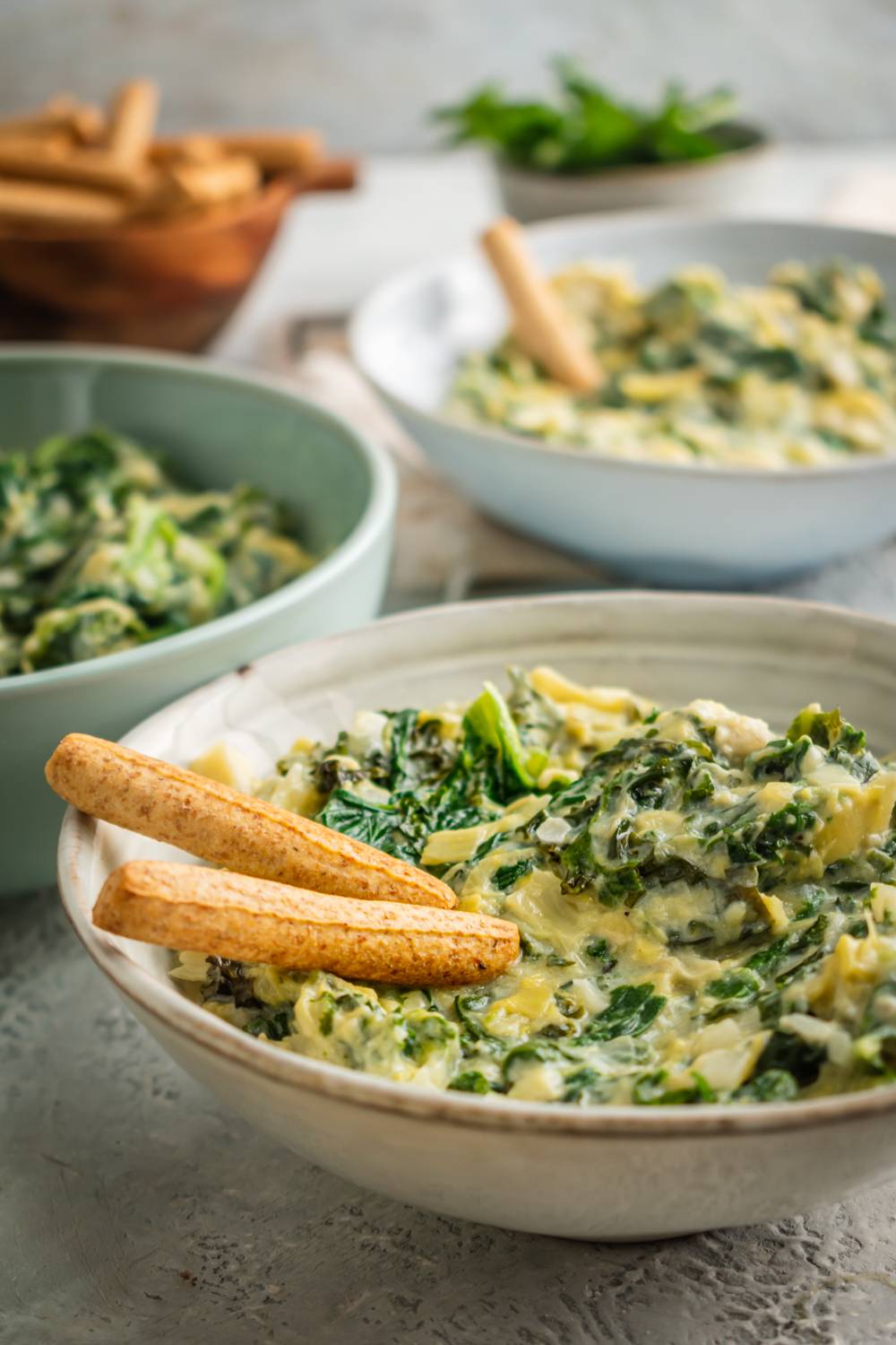 A creamy spinach, artichoke, and kale dip served in a bowl with crispy breadsticks for dipping, surrounded by additional bowls of dip.