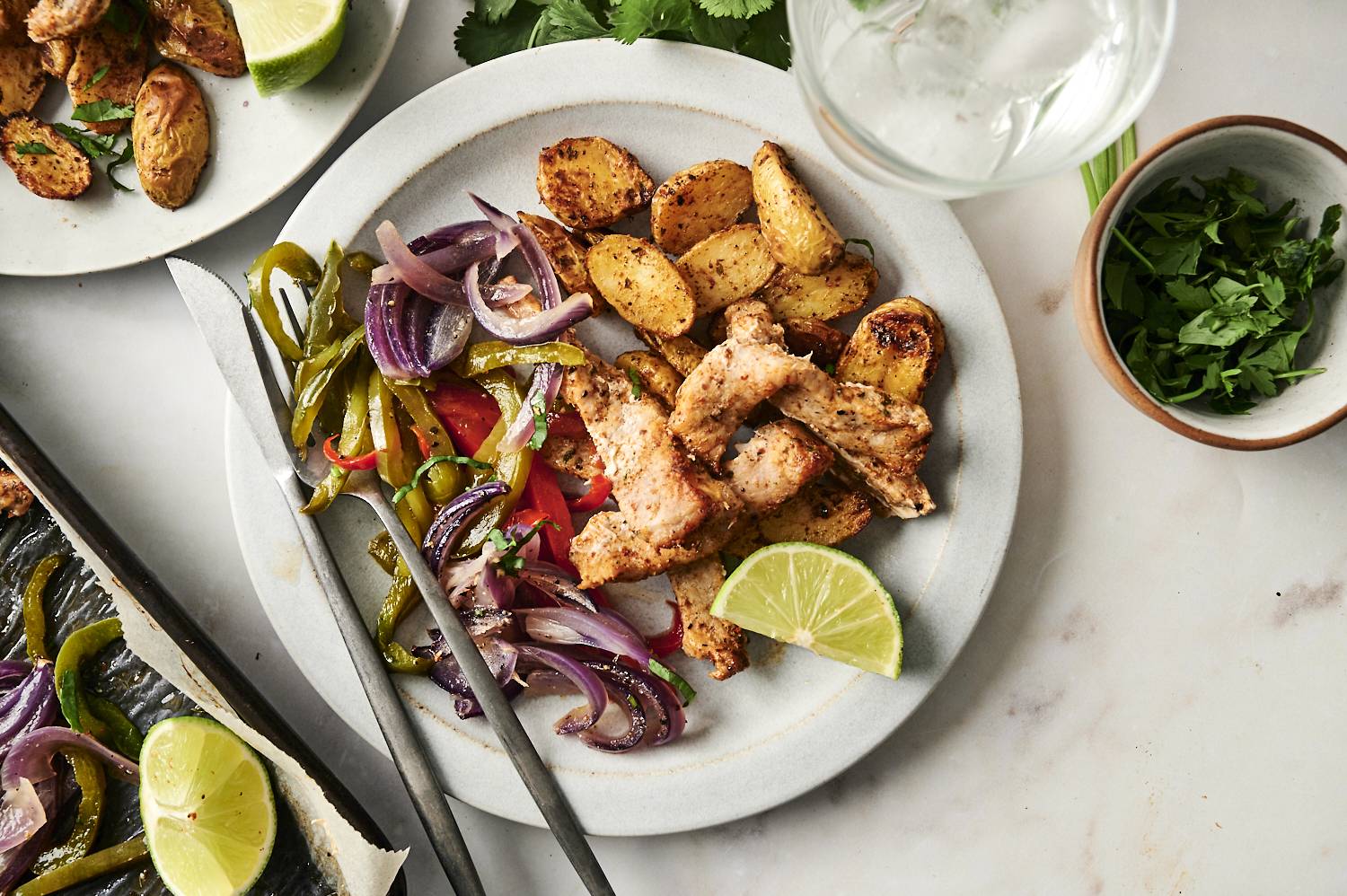 A plated meal of seasoned pork chops, roasted potatoes, and sautéed bell peppers with red onions, garnished with fresh cilantro and a lime wedge.