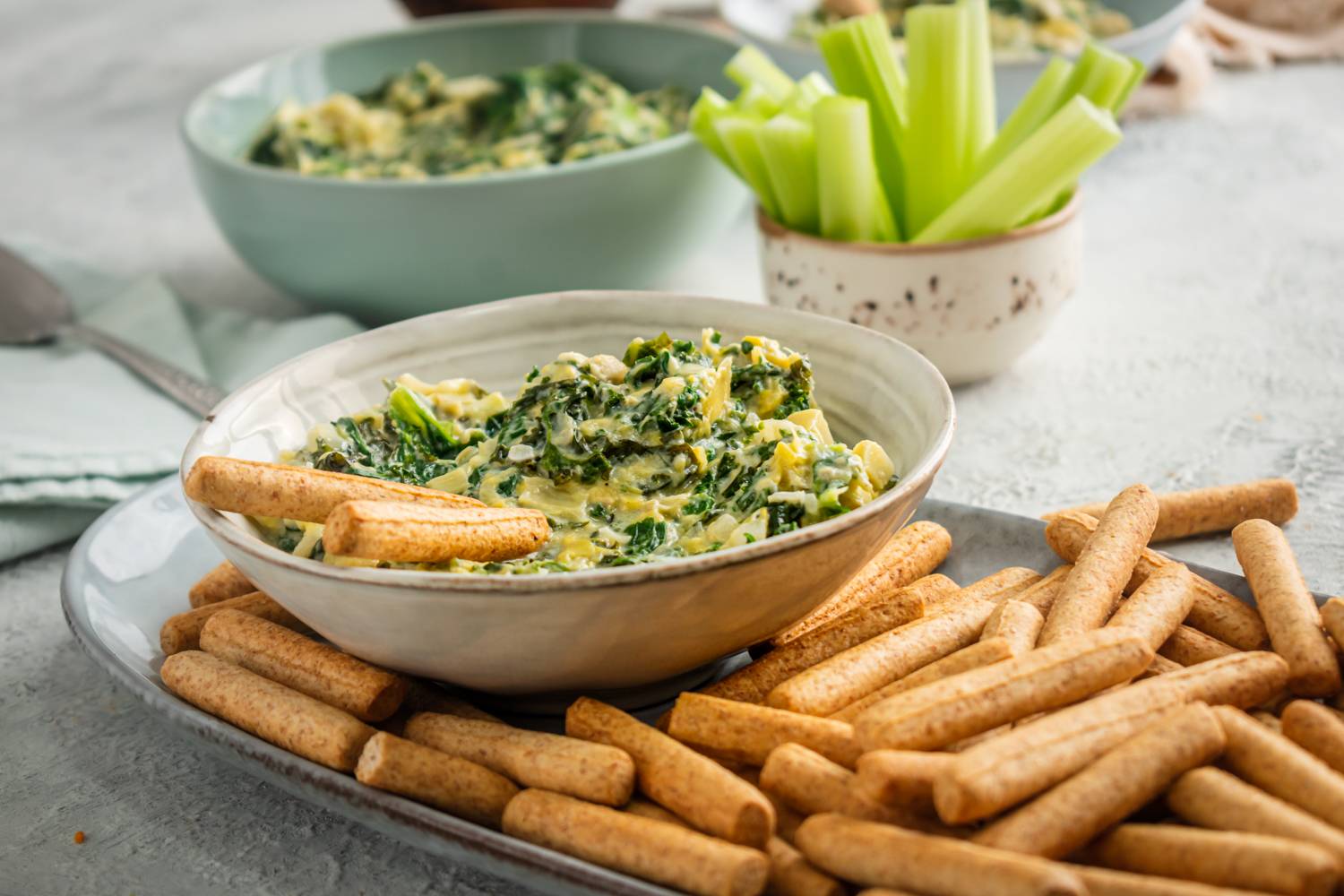 A bowl of creamy spinach, artichoke, and kale dip served with crunchy breadsticks and a side of fresh celery for dipping.