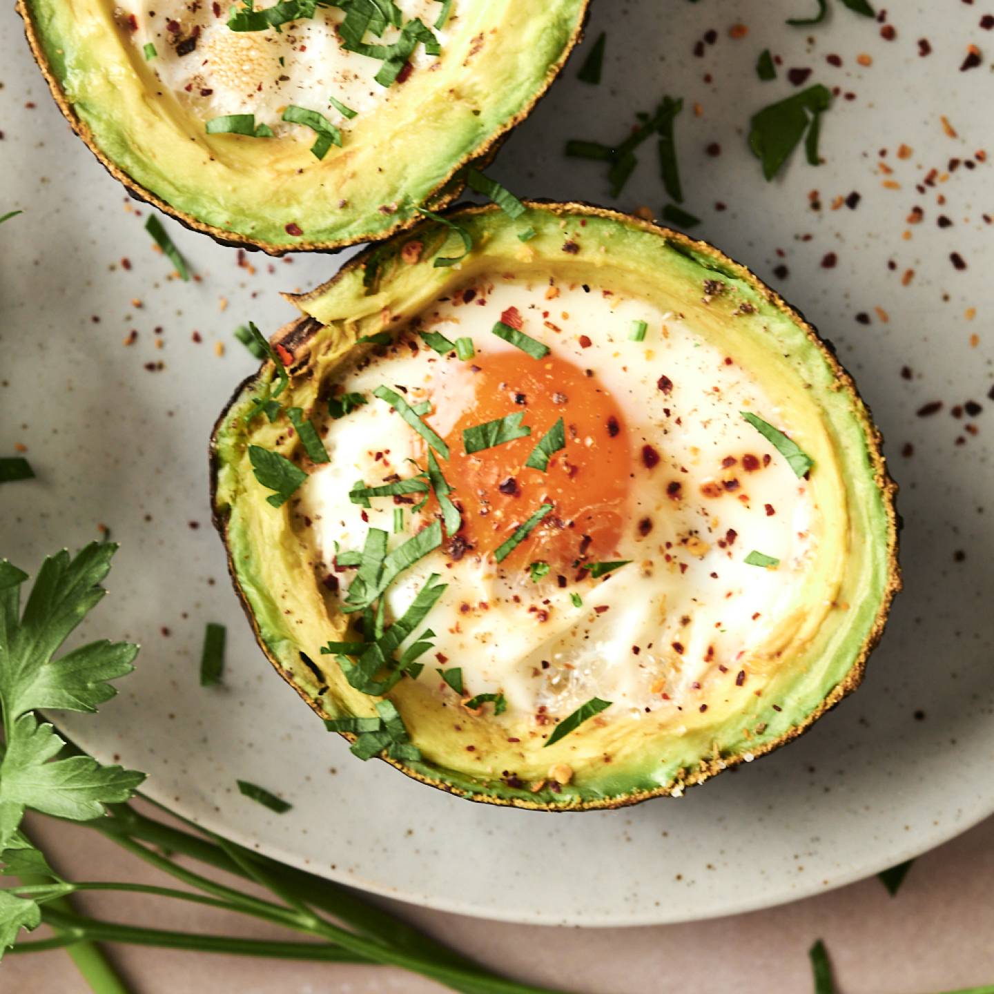Two avocado halves baked with eggs, sprinkled with red pepper flakes and fresh chopped parsley, served on a neutral plate with parsley garnish on the side.