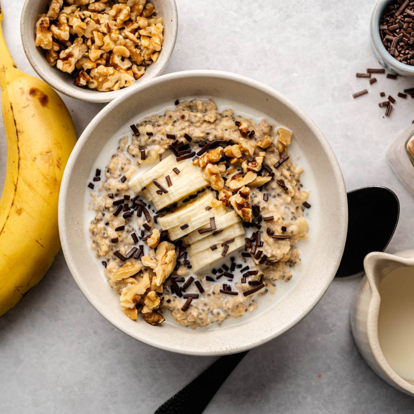 A creamy oatmeal bowl topped with banana slices, walnuts, and chocolate sprinkles. Nearby are ripe bananas, walnuts, chocolate sprinkles, and milk. Warm, inviting tone.