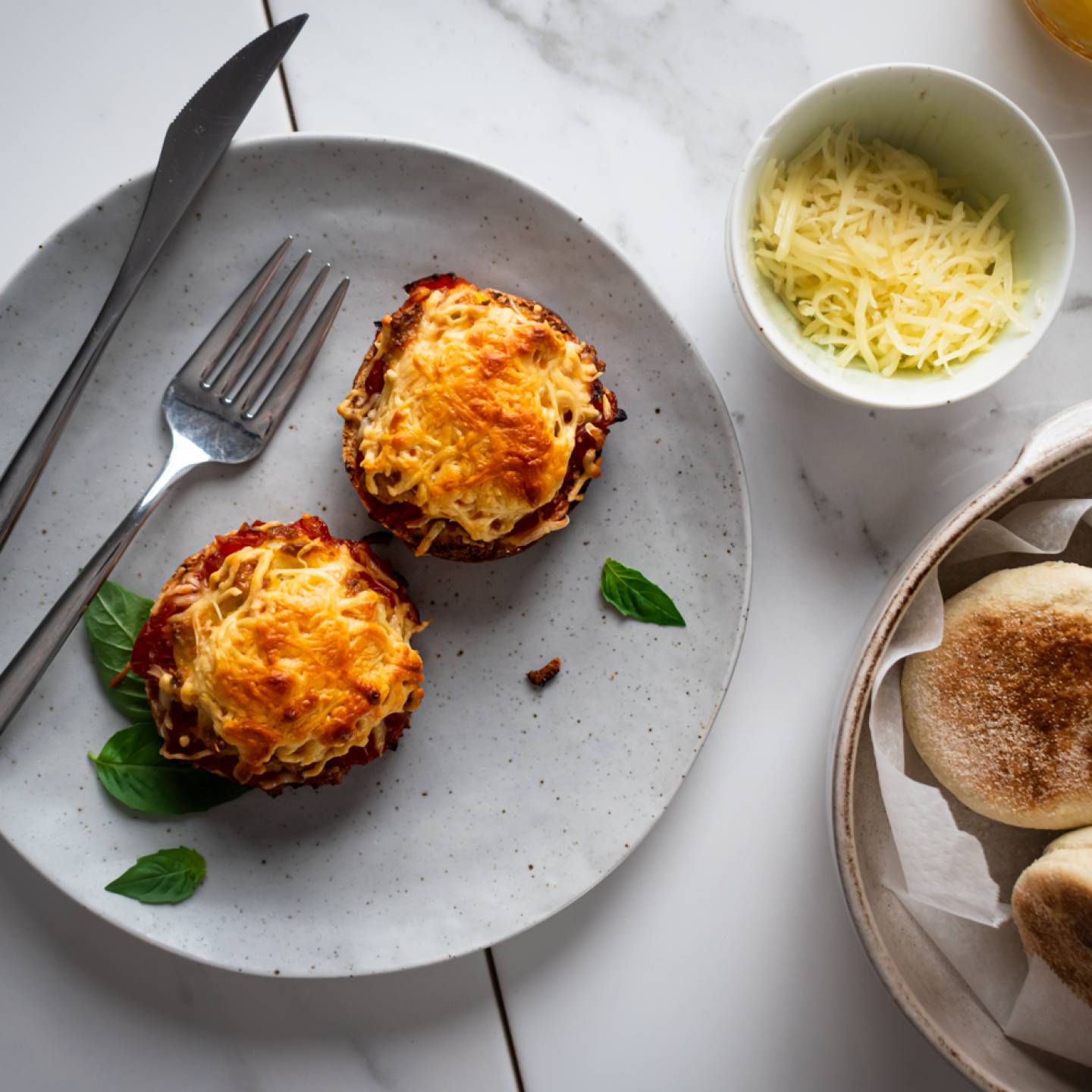 Breakfast English muffin pizzas with eggs, tomato sauce, basil, and melted cheese on a plate with English muffins on the side.