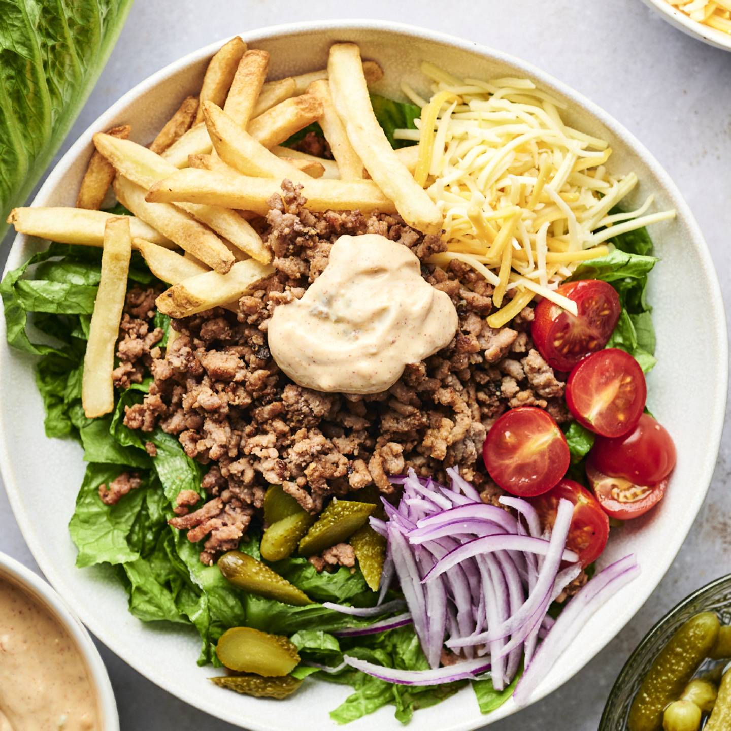 Burger bowls with seasoned ground beef, french fries, lettuce, tomatoes, red onion, pickles, and special sauce in a bowl.