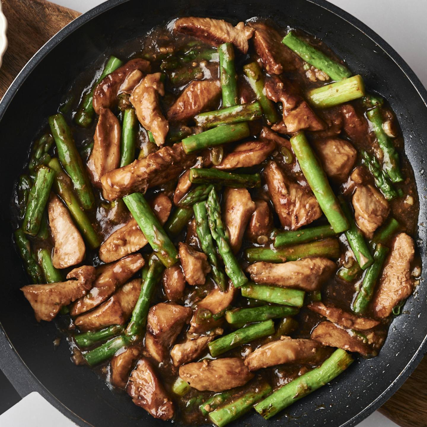 Stir-fried chicken and asparagus in a dark sauce, garnished with chopped green onions in a bowl on the side.