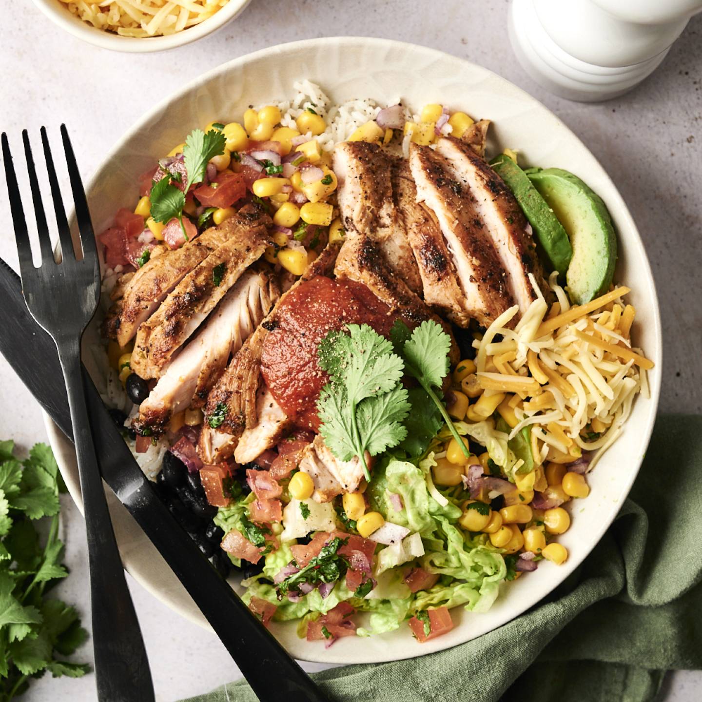 A colorful bowl featuring grilled chicken slices, corn, cheese, lettuce, and salsa, garnished with cilantro and avocado.