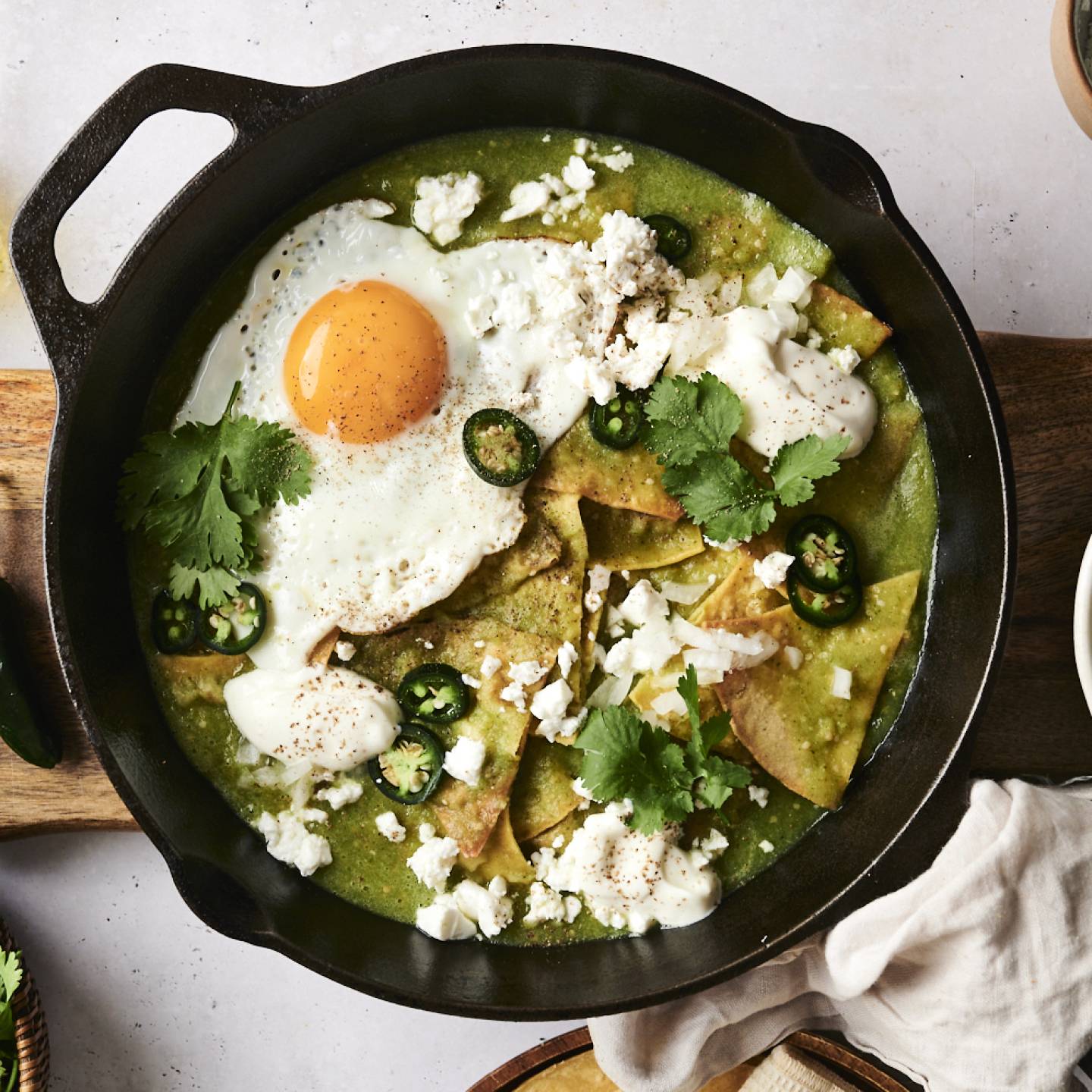 Chilaquiles verdes in a cast iron pan, topped with a fried egg, crumbled cheese, sour cream, jalapeño slices, and fresh cilantro. The pan is placed on a wooden board, surrounded by bowls of chopped onions, cheese, and fresh cilantro. A jalapeño and a bottle of oil are also nearby.