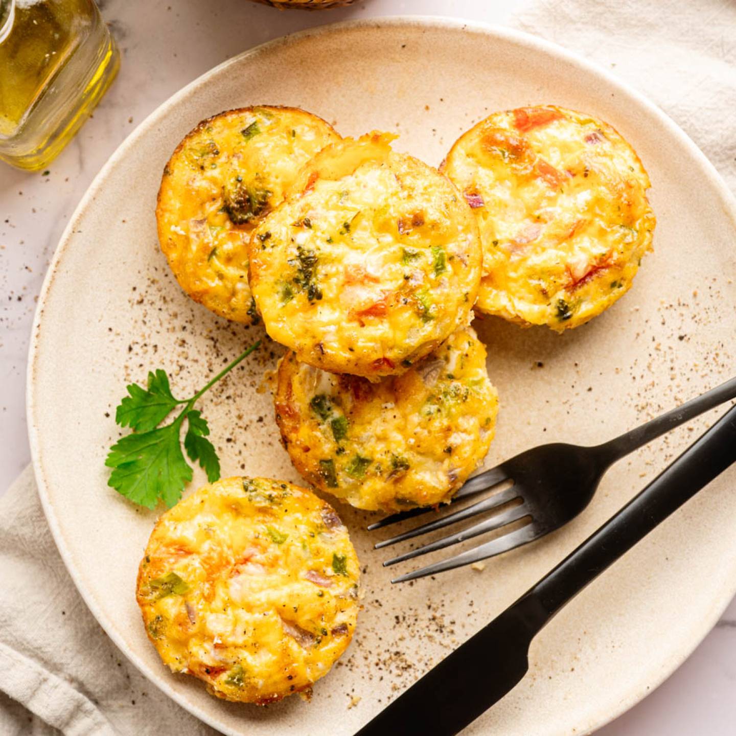 Plate of five colorful egg muffins with veggies, garnished with a parsley leaf. Accompanied by a black fork, napkin, and nearby olive oil. Inviting and fresh.