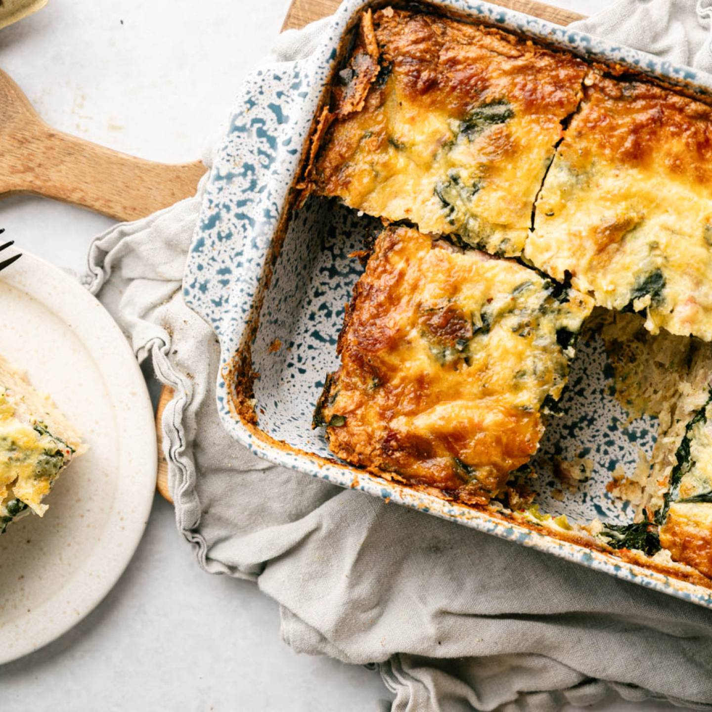 Golden-brown spinach and cheese casserole in a speckled baking dish, partially cut. A single slice on a white plate with a fork, set on a wooden board.