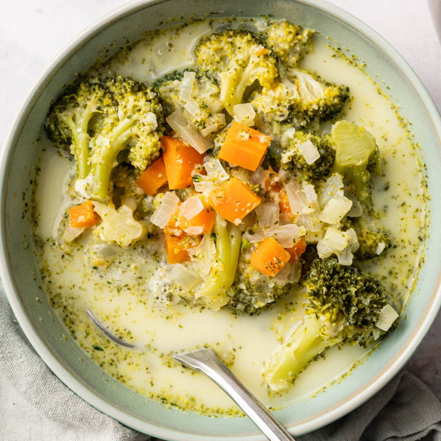 A bowl of broccoli cheddar soup with visible broccoli florets, diced carrots, and onion chunks in a creamy broth. A spoon rests on the bowl's edge. The soup sits on a light-colored cloth, conveying a comforting, homemade feel.