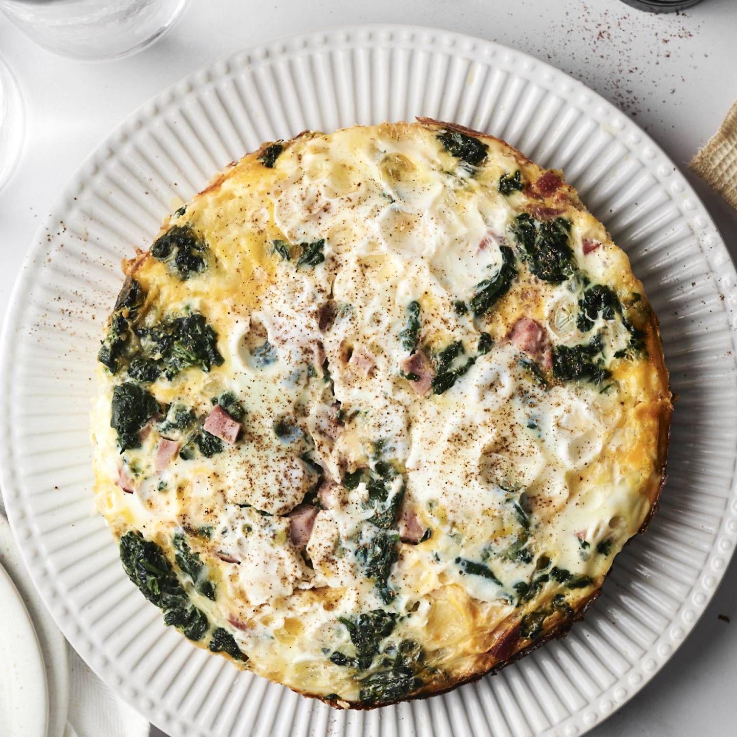A close-up of a slice of breakfast casserole with layers of hash browns, eggs, ham, spinach, and cheese on a plate, with a fork resting beside it.