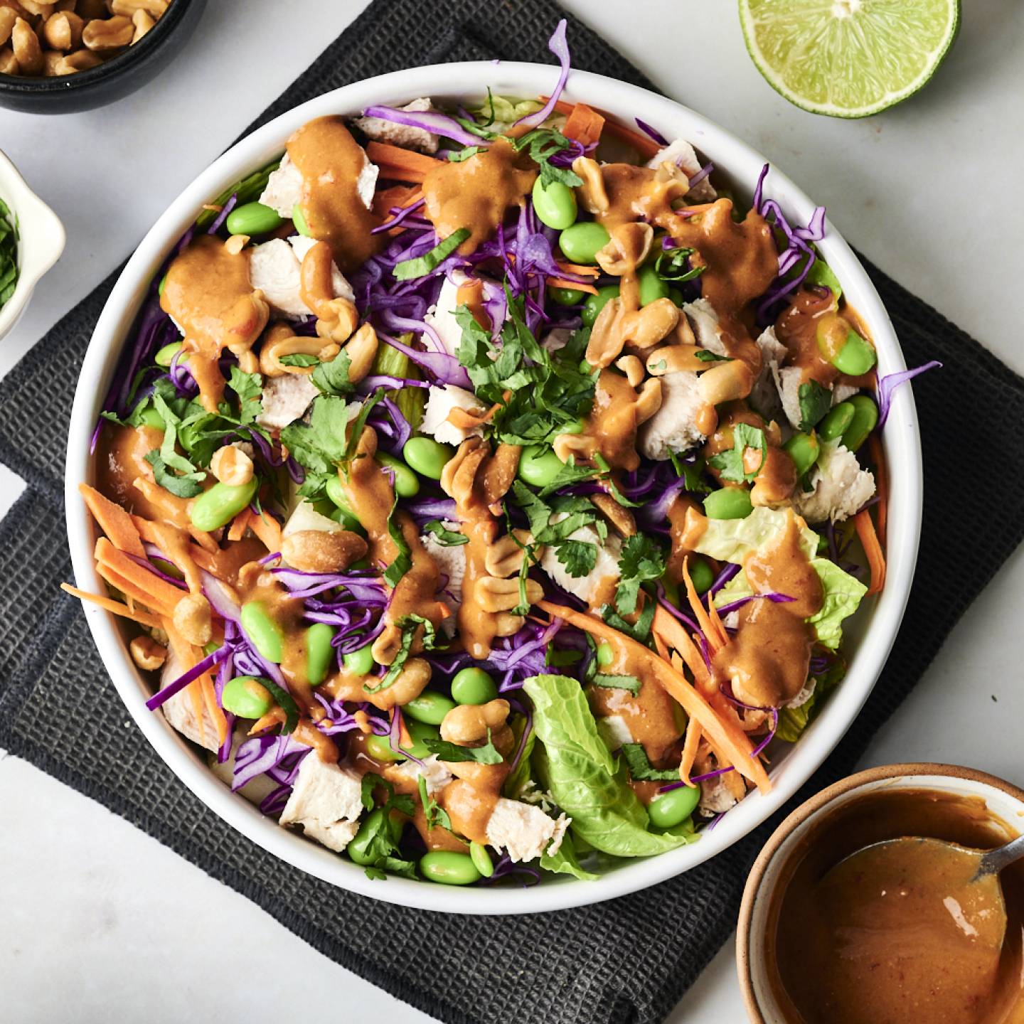 A vibrant salad with purple cabbage, carrots, edamame, chicken, and cilantro, drizzled with peanut sauce and topped with cashews.