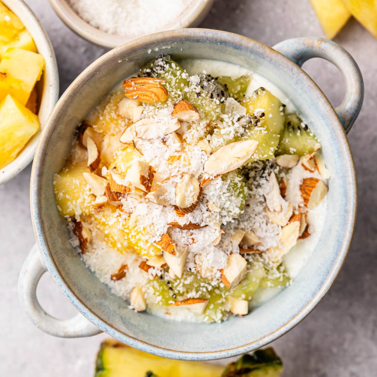 A bowl of yogurt topped with chopped almonds, shredded coconut, kiwi slices, and pineapple chunks. Surrounding it are bowls of pineapple and kiwi on a gray surface.