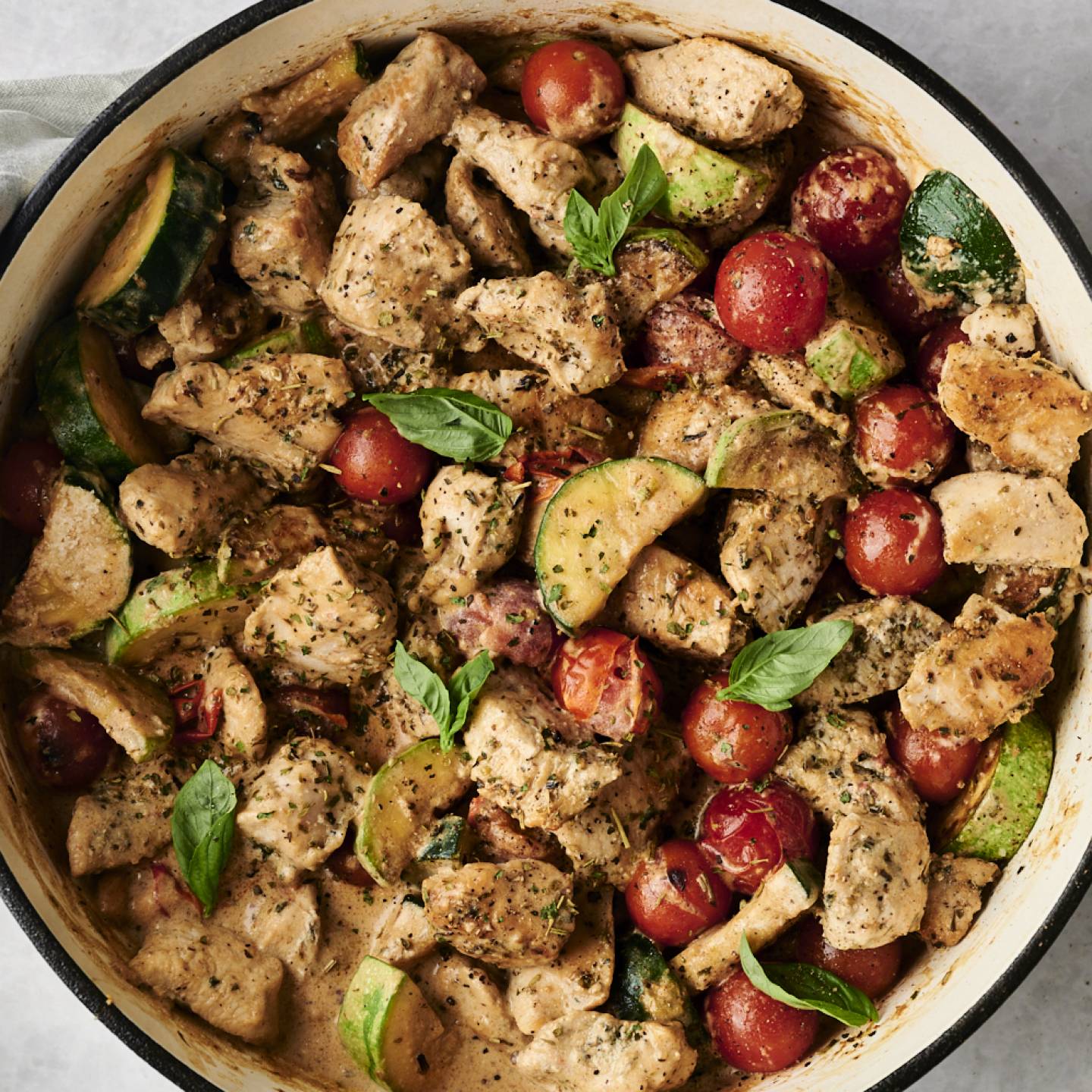 Top-down view of a creamy Tuscan chicken skillet filled with chunks of seasoned chicken, cherry tomatoes, zucchini slices, and fresh basil in a creamy herb sauce. A gray napkin and small bowl of Italian seasoning rest nearby.
