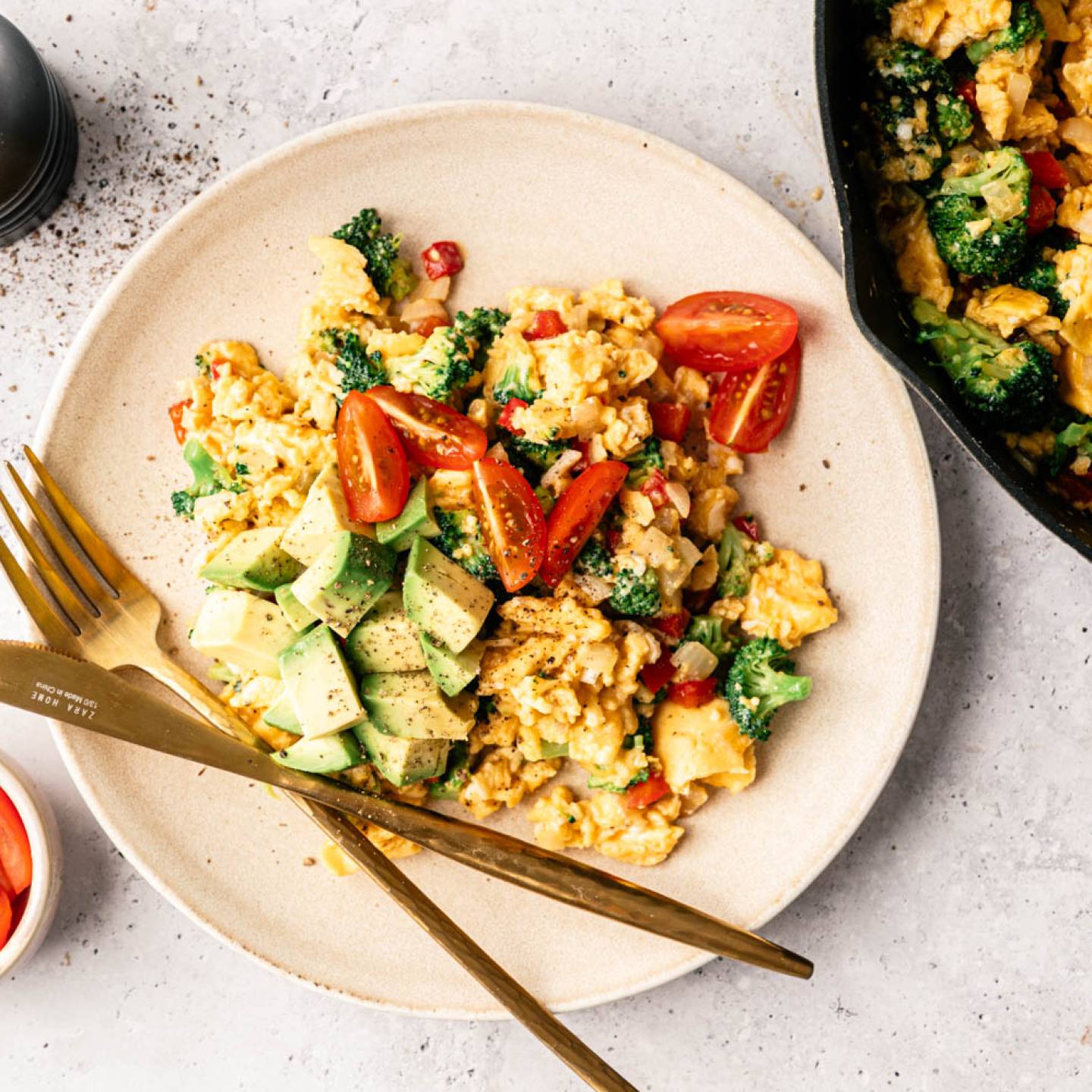 A plate of scrambled eggs mixed with broccoli and red peppers, topped with avocado and cherry tomatoes. A fork and knife rest beside it. A bowl of tomatoes and a skillet are nearby. The setting is bright and appetizing.