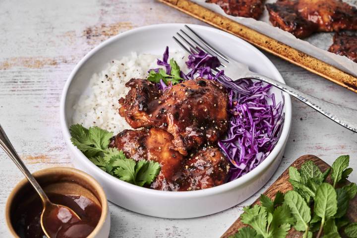 A white bowl filled with baked BBQ chicken thighs glazed in a rich sauce, served alongside fluffy white rice, shredded purple cabbage, and fresh cilantro, with a fork resting on the side.
