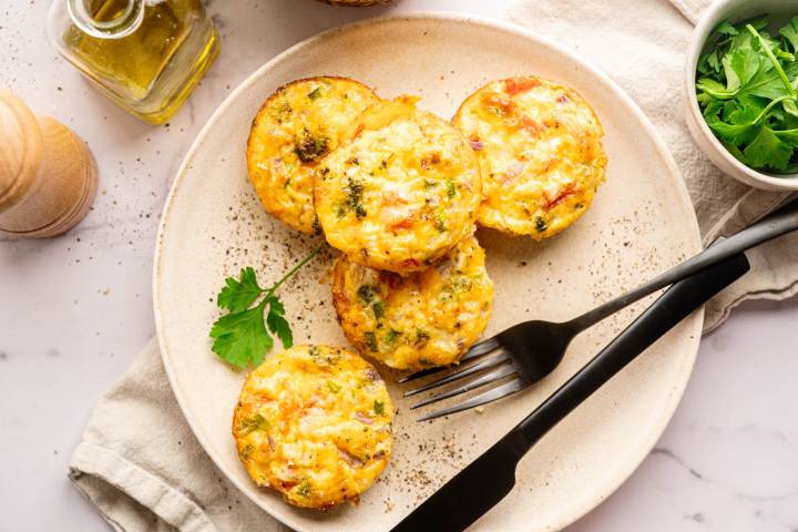 Plate of five colorful egg muffins with veggies, garnished with a parsley leaf. Accompanied by a black fork, napkin, and nearby olive oil. Inviting and fresh.