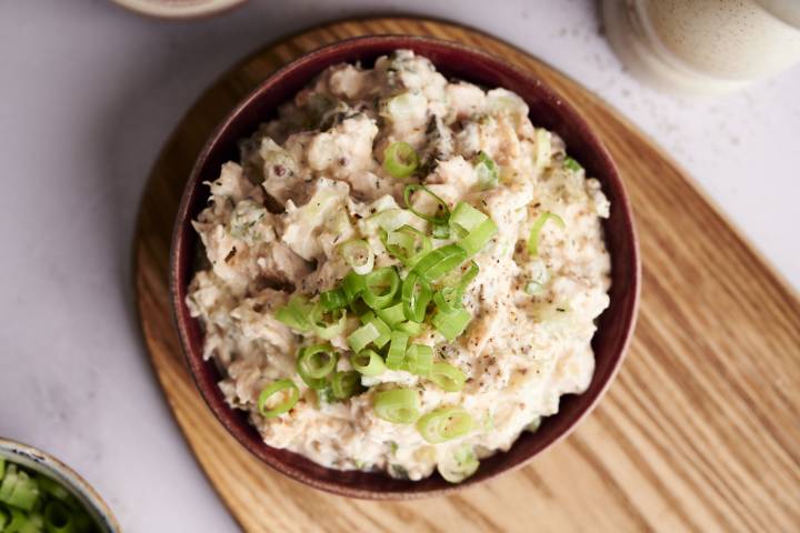 A top-down shot of a bowl of dill pickle salmon salad, garnished with sliced green onions and cracked black pepper, on a wooden cutting board.