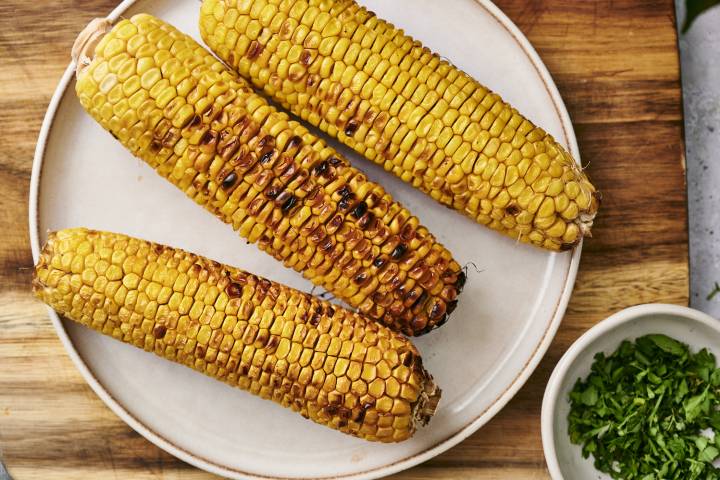 Three grilled corn cobs with golden kernels sit on a white plate beside a small bowl of chopped green herbs.