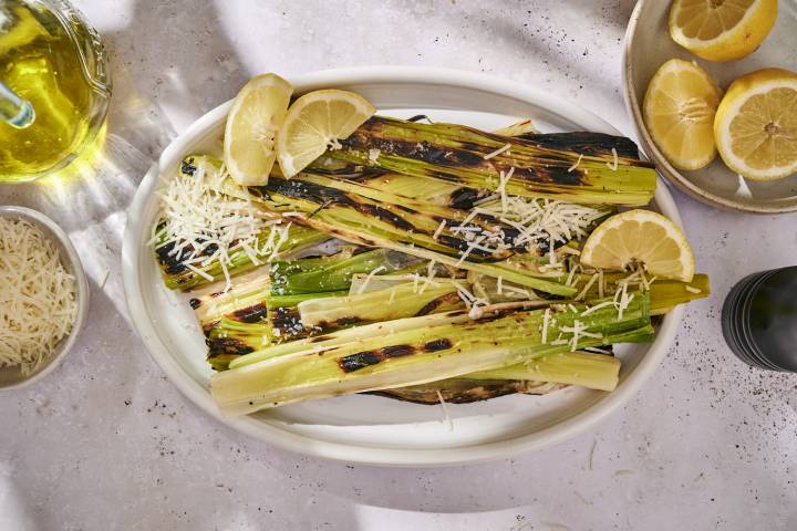 Charred grilled leeks served on a white platter with lemon wedges and a sprinkle of shredded Parmesan cheese.