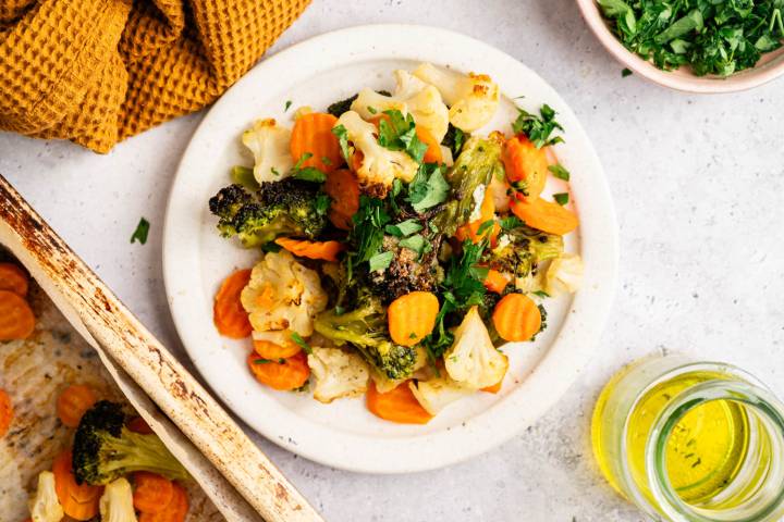A white plate with roasted broccoli, cauliflower, and sliced carrots topped with fresh parsley. Surrounded by a yellow cloth, a dish of herbs, and olive oil.