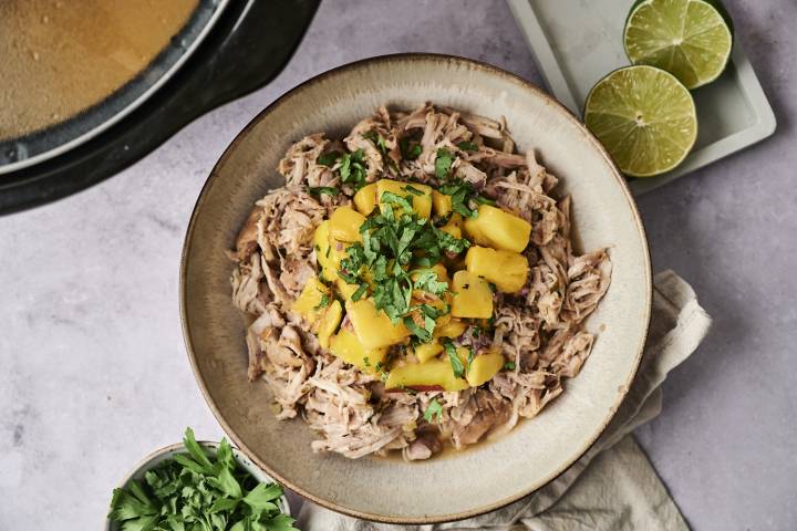 A bowl of shredded meat topped with diced mango and fresh cilantro, surrounded by lime halves and fresh herbs.