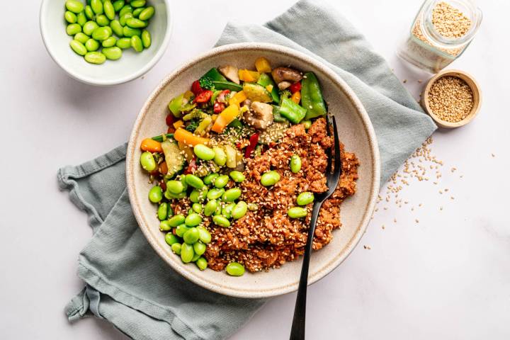 A bowl of colorful vegan stir-fry with edamame, quinoa, and vegetables on a gray cloth. Nearby are scattered sesame seeds and a small bowl of edamame.