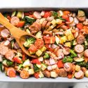 One pan sausage, potato, and veggie in a baking dish with a wooden spoon.