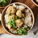 A bowl of white rice topped with Thai curry chicken meatballs, coated in a creamy sauce and garnished with fresh cilantro and sliced green onions.