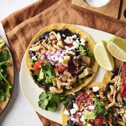 A  plate of tostadas, topped with black beans, shredded chicken, fresh lettuce, tomato, red onion, avocado, cilantro, and crumbled cheese. Lime wedges are placed on the side for a fresh squeeze of citrus.