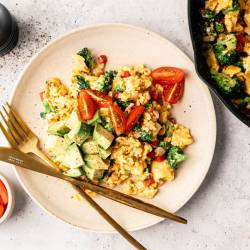 A plate of scrambled eggs mixed with broccoli and red peppers, topped with avocado and cherry tomatoes. A fork and knife rest beside it. A bowl of tomatoes and a skillet are nearby. The setting is bright and appetizing.