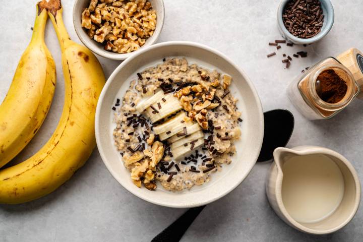 A creamy oatmeal bowl topped with banana slices, walnuts, and chocolate sprinkles. Nearby are ripe bananas, walnuts, chocolate sprinkles, and milk. Warm, inviting tone.