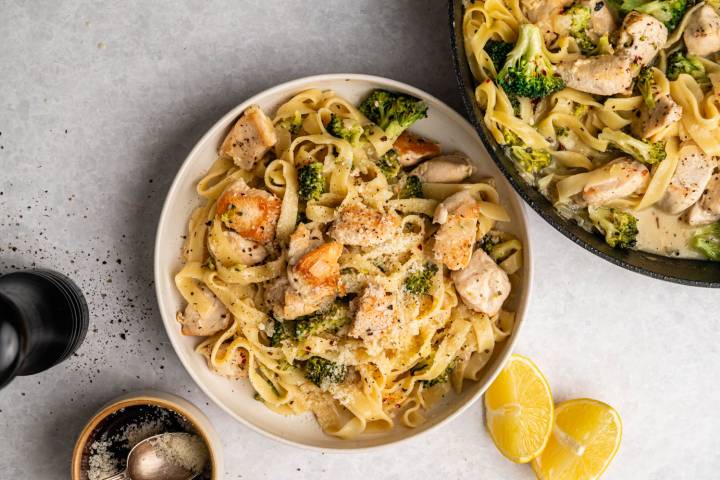 Plate of creamy chicken and broccoli pasta topped with parmesan, next to a similar skillet. Lemon wedges and pepper grinder add fresh vibes.