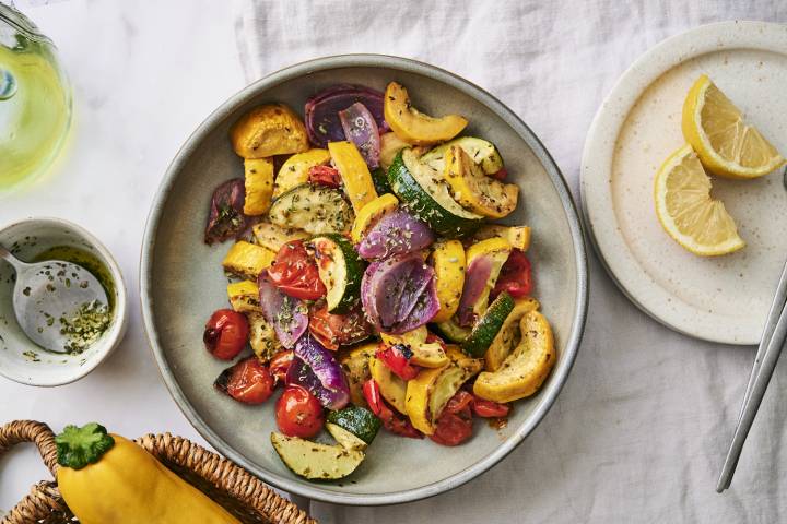 A colorful medley of roasted zucchini, yellow squash, cherry tomatoes, and red onion served with lemon on a light table setting.