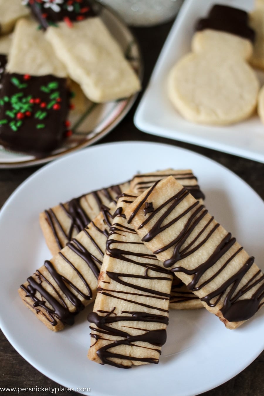 Chocolate Dipped Shortbread Cookies are a simple, from-scratch, shortbread cookie dipped in dark chocolate that's perfect for dunking in milk, coffee, hot chocolate...Santa won't be disappointed with these shortbread Christmas cookies! | www.persnicketyplates.com #christmascookies #shortbread #easyrecipe #baking #cookies #chocolate #sprinkles white plate of shortbread cookies drizzled with chocolate