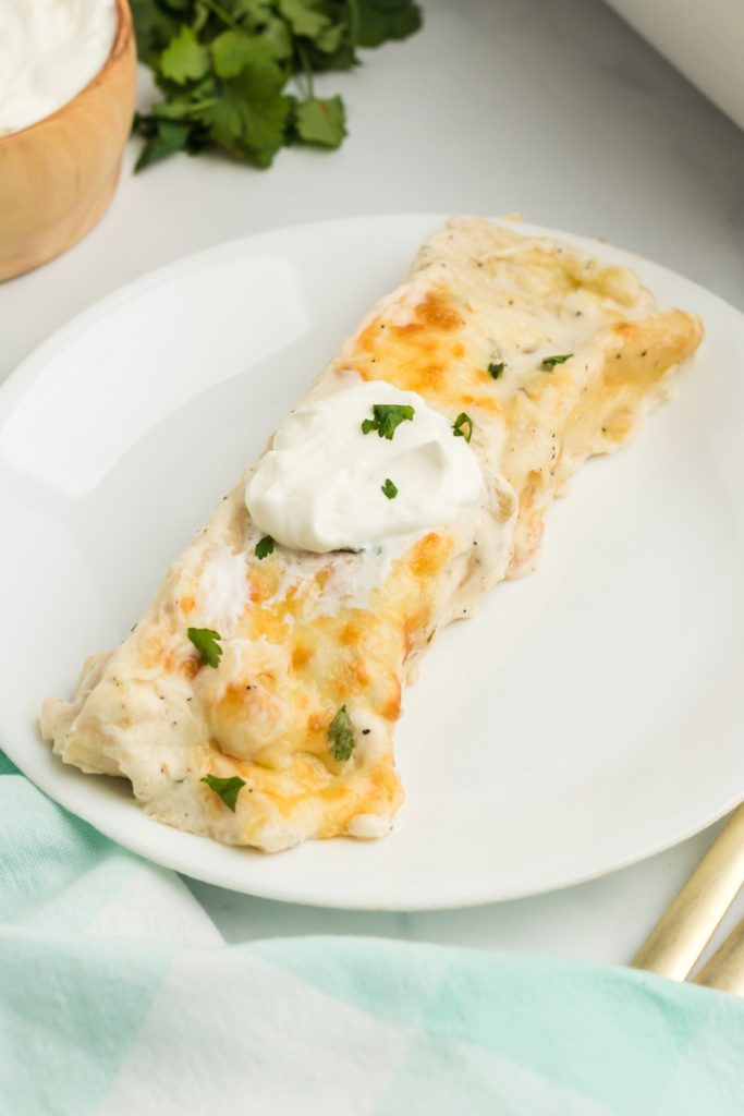 a single enchilada on a white plate topped with sour cream and cilantro.