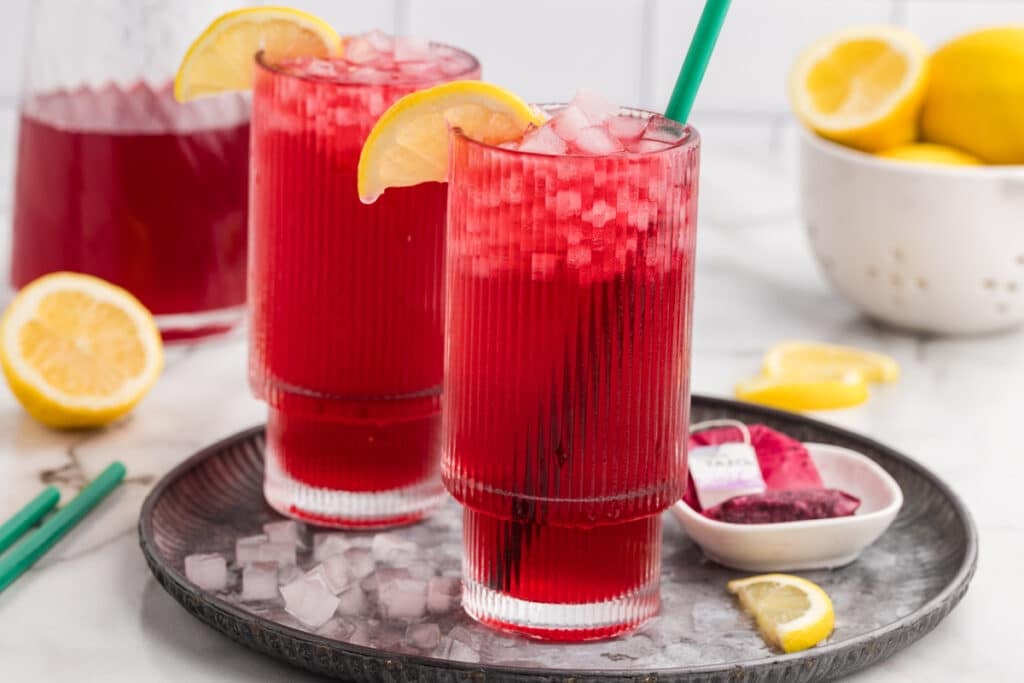 Two ribbed glasses of iced passion tea lemonade on a tray with lemon slices and tea bags.