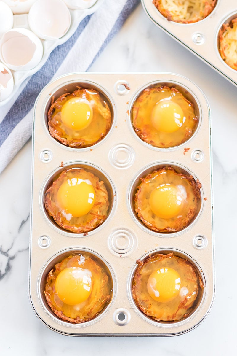 Bird's Nest Breakfast Cups are a fun way to eat eggs and hash browns for breakfast. These baked egg cups combine hash browns and cheese to form a "nest" and are topped with an egg. | www.persnicketyplates.com eggs cracked into hash brown cups in a muffin tin