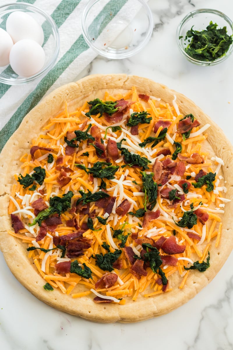 Bacon & spinach breakfast pizza is a filling and unique way to start your day. Pizza for breakfast is always a good idea! | www.persnicketyplates.com #pizza #breakfast #breakfastpizza #easyrecipe cheese, spinach, and bacon sprinkled on a pizza crust
