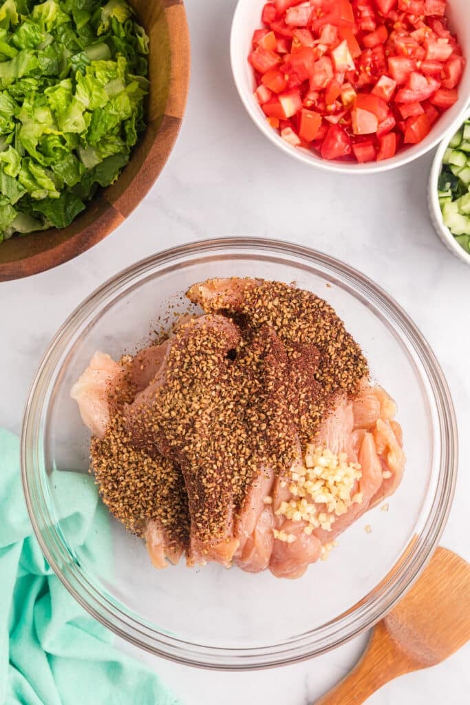seasonings sprinkled on raw chicken in mixing bowl.